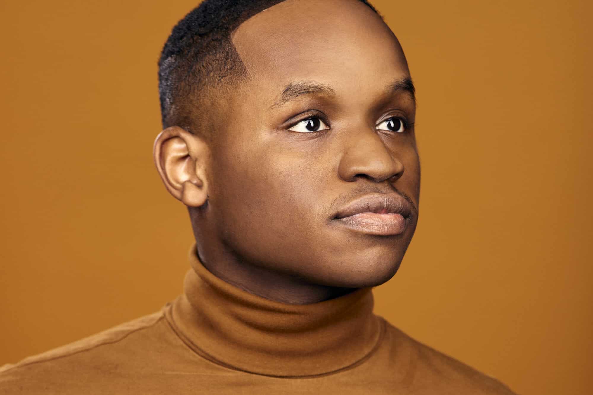 NYC Dancer Joshua Dawson's headshot. He's wearing a brown cowl neck stood against a brown background