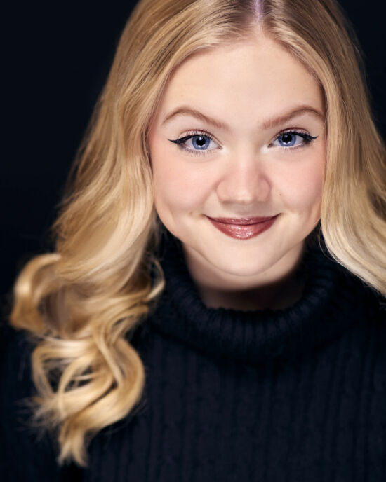 Headshots of Abigail Douglas, a blonde hair, blue eyed Child Actor based out of New York City. She's wearing a black polo jumper, photographed against a black savage universal paper background
